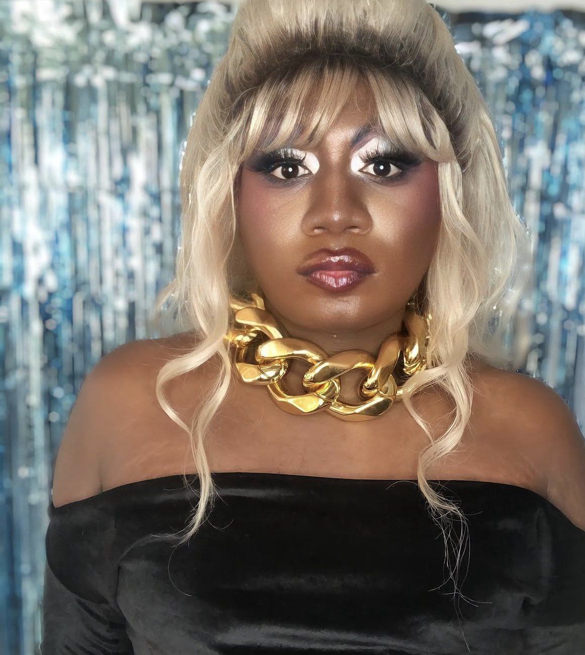 Yshee Black in a blonde wig and gold chain against silver tinsel